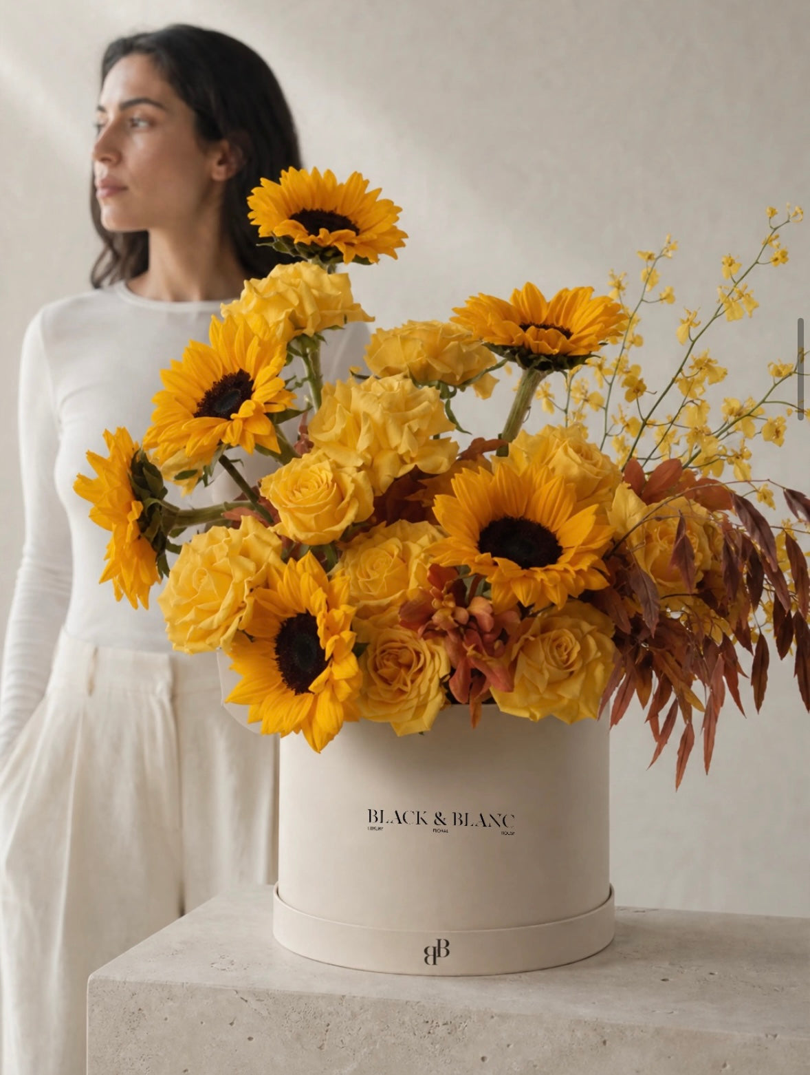 A woman in white beside a Black & Blanc box of sunflowers, golden roses and yellow oncidium orchid