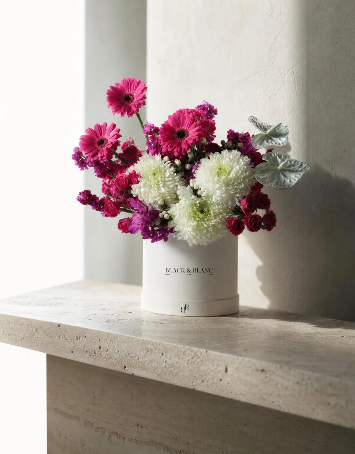 Pink gerbera, spray roses and white chrysanthemum in a Black & Blanc box on a travertine ledge