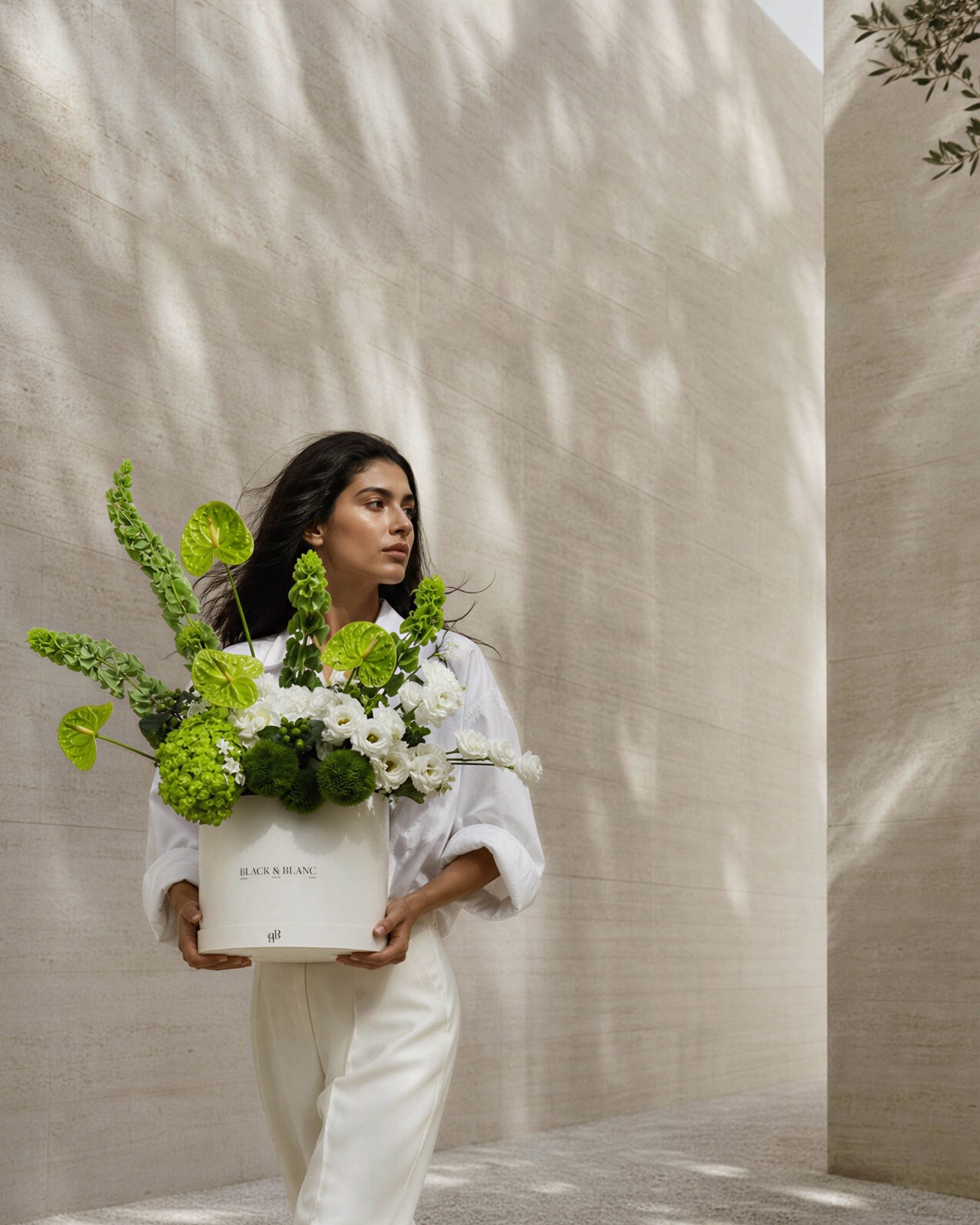 A woman in white carries a Black & Blanc box of green anthurium and white lisianthus along a sunlit travertine wall