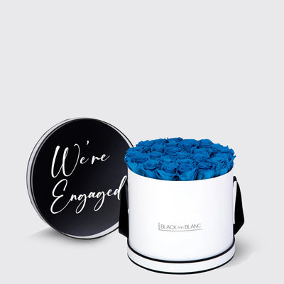 Cylindrical white box of vibrant blue infinity roses with a black We’re Engaged lid